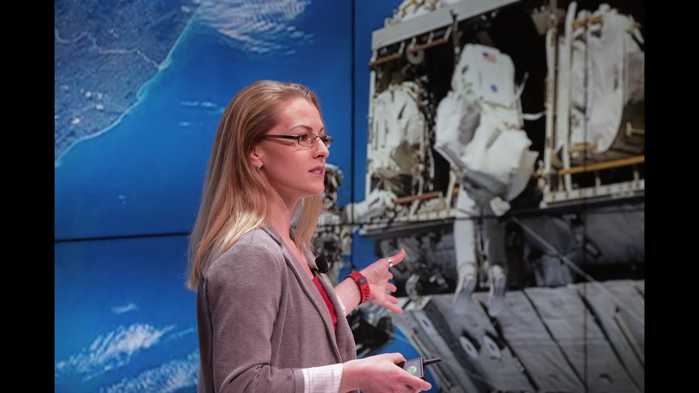 A woman wearing glasses gives a presentation in front of a large screen.